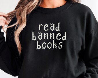 May include: Black sweatshirt with the text "read banned books" in white gothic font. The sweatshirt is a crew neck style and is being worn with denim shorts. The word "BLACK" is in a black circle in the bottom right corner.