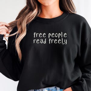 Może przedstawiać: Czarna bluza z białym napisem "free people read freely" w gotyckiej czcionce. Top z długim rękawem jest połączony z dżinsowymi szortami. Czarne koło ze słowem "BLACK" znajduje się w prawym dolnym rogu.