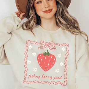 May include: A cream-colored sweatshirt with a pink strawberry graphic and the text "feeling berry good" in a pink frame.
