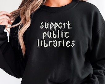 May include: Black sweatshirt with the text "support public libraries" in a white gothic font. The sweatshirt is a crew neck style and is being worn with denim shorts. The word "BLACK" is in a circle in the bottom right corner.