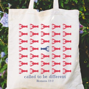 May include: Cream-colored tote bag with a repeating pattern of red lobsters, with one blue lobster. The text "called to be different" and "Romans 12:2" are printed in blue. The bag has white handles.