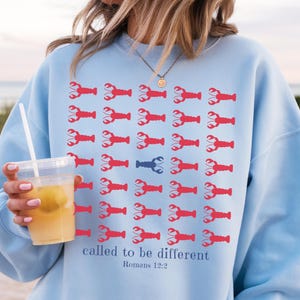 May include: A light blue crewneck sweatshirt featuring a grid of red lobster icons with one single blue lobster in the center. Below the grid, the text reads "called to be different" followed by "Romans 12:2" in a dark blue serif font.
