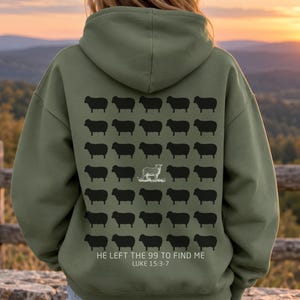 May include: A military green hooded sweatshirt featuring a back graphic of forty-nine black sheep silhouettes arranged in a grid, with one white sheep outline in the center. Text below reads: HE LEFT THE 99 TO FIND ME LUKE 15:3-7.
