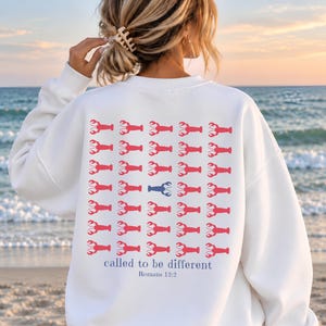 May include: A white long-sleeve sweatshirt featuring a grid of red lobster graphics on the back. One blue lobster is positioned in the center of the grid. Below the lobsters, the text reads, "called to be different" and "Romans 12:2".