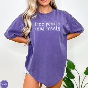 May include: A purple t-shirt with the text "free people read freely" in white script. The shirt has short sleeves and a relaxed fit. The shirt is a solid grape color.