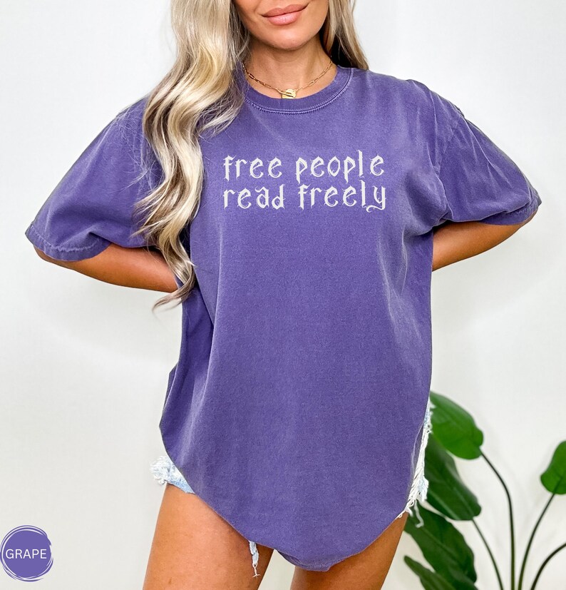 May include: A purple t-shirt with the text "free people read freely" in white script. The shirt has short sleeves and a relaxed fit. The shirt is a solid grape color.
