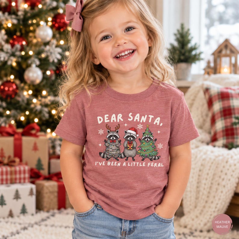 Puede incluir: Una camiseta de manga corta en color malva jaspeado con un gr&aacute;fico de tres mapaches y el texto "DEAR SANTA, I'VE BEEN A LITTLE FERAL". Un mapache lleva astas de reno, otro un gorro de Pap&aacute; Noel y el tercero est&aacute; envuelto en luces de Navidad.