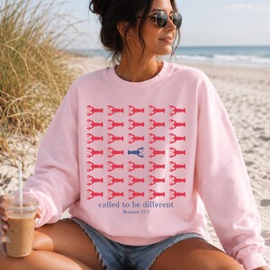 May include: A light pink crewneck sweatshirt featuring a repeating grid pattern of red lobster icons, with one blue lobster in the center. Below the grid, the text reads "called to be different" in blue, followed by "Romans 12:2" in smaller blue text.