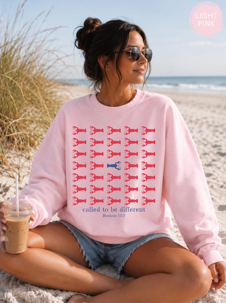 May include: A light pink crewneck sweatshirt featuring a repeating grid pattern of red lobster icons, with one blue lobster in the center. Below the grid, the text reads "called to be different" in blue, followed by "Romans 12:2" in smaller blue text.