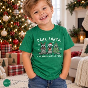 Puede incluir: Una camiseta verde de manga corta con un gr&aacute;fico de tres mapaches con decoraciones navide&ntilde;as y el texto "DEAR SANTA, I'VE BEEN A LITTLE FERAL" impreso en blanco. El dise&ntilde;o incluye copos de nieve y elementos festivos navide&ntilde;os.