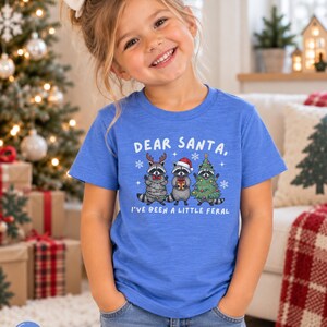 Puede incluir: Una camiseta azul jaspeado de manga corta con un gr&aacute;fico de tres mapaches y el texto "DEAR SANTA, I'VE BEEN A LITTLE FERAL". Un mapache lleva astas de reno, otro un gorro de Pap&aacute; Noel y el tercero est&aacute; envuelto en un &aacute;rbol de Navidad.
