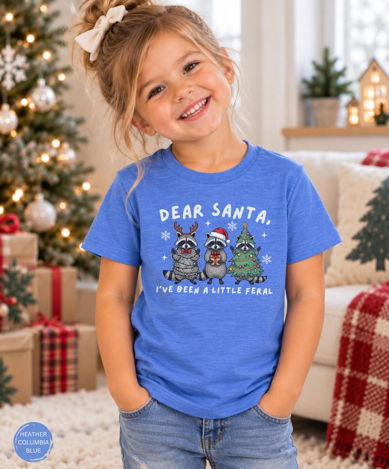 Puede incluir: Una camiseta azul jaspeado de manga corta con un gr&aacute;fico de tres mapaches y el texto "DEAR SANTA, I'VE BEEN A LITTLE FERAL". Un mapache lleva astas de reno, otro un gorro de Pap&aacute; Noel y el tercero est&aacute; envuelto en un &aacute;rbol de Navidad.