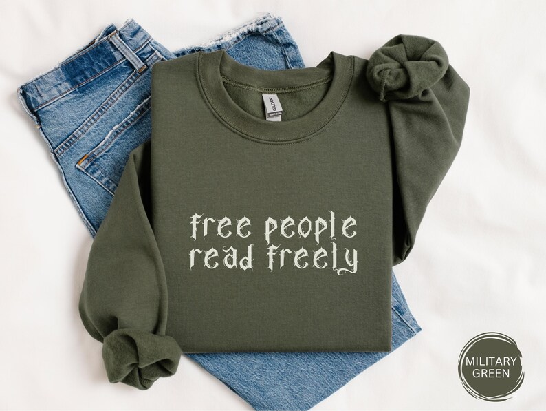 Może przedstawiać: Oliwkowozielona bluza z napisem "free people read freely" gotycką czcionką. Bluza jest złożona na parze niebieskich dżins&oacute;w. Kolor bluzy oznaczony jest jako "Military Green".