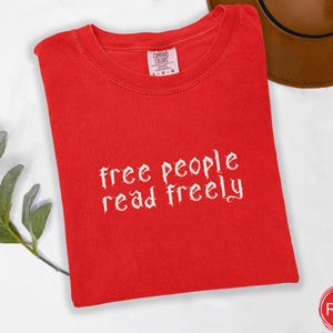 May include: A bright red t-shirt with the text "free people read freely" in white script. The shirt is folded neatly, with a tag visible at the top. A brown hat is partially visible in the background.