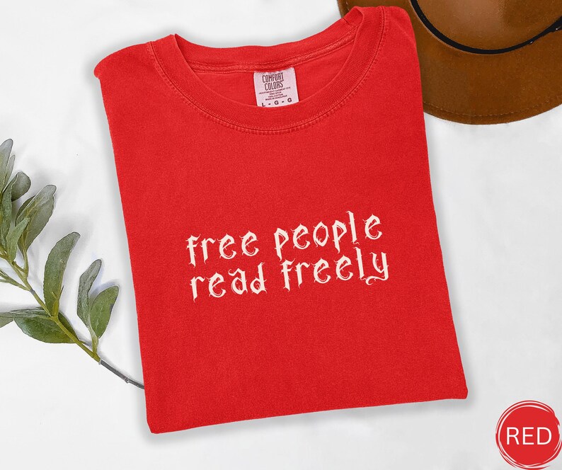 May include: A bright red t-shirt with the text "free people read freely" in white script. The shirt is folded neatly, with a tag visible at the top. A brown hat is partially visible in the background.