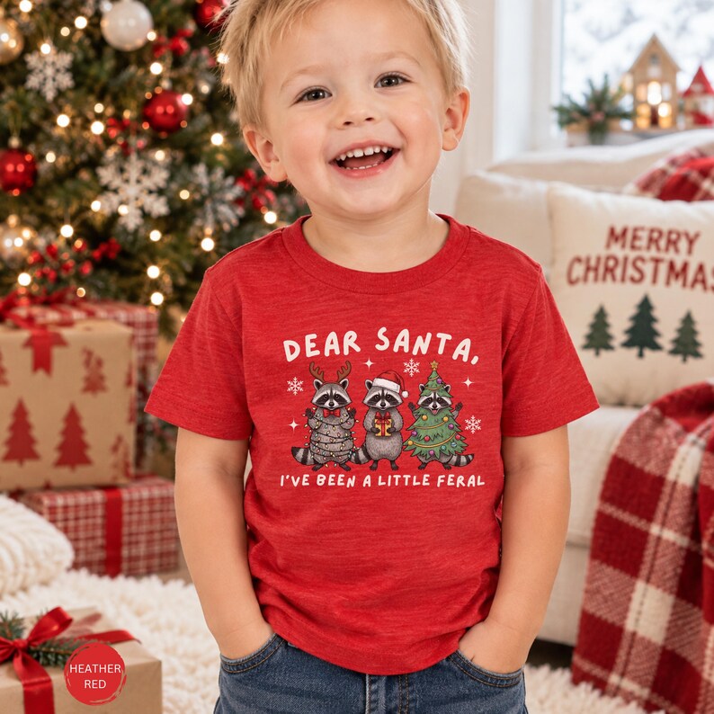 Puede incluir: Una camiseta de manga corta en color rojo jaspeado con un gr&aacute;fico de tres mapaches y el texto "DEAR SANTA, I'VE BEEN A LITTLE FERAL". Un mapache lleva astas de reno, otro un gorro de Pap&aacute; Noel y el tercero est&aacute; decorado como un &aacute;rbol de Navidad.