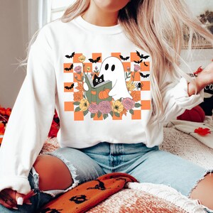 May include: White sweatshirt featuring a Halloween-themed graphic. The design includes a ghost reading a book with a black cat, surrounded by flowers, pumpkins, and bats, set against an orange and white checkered background.