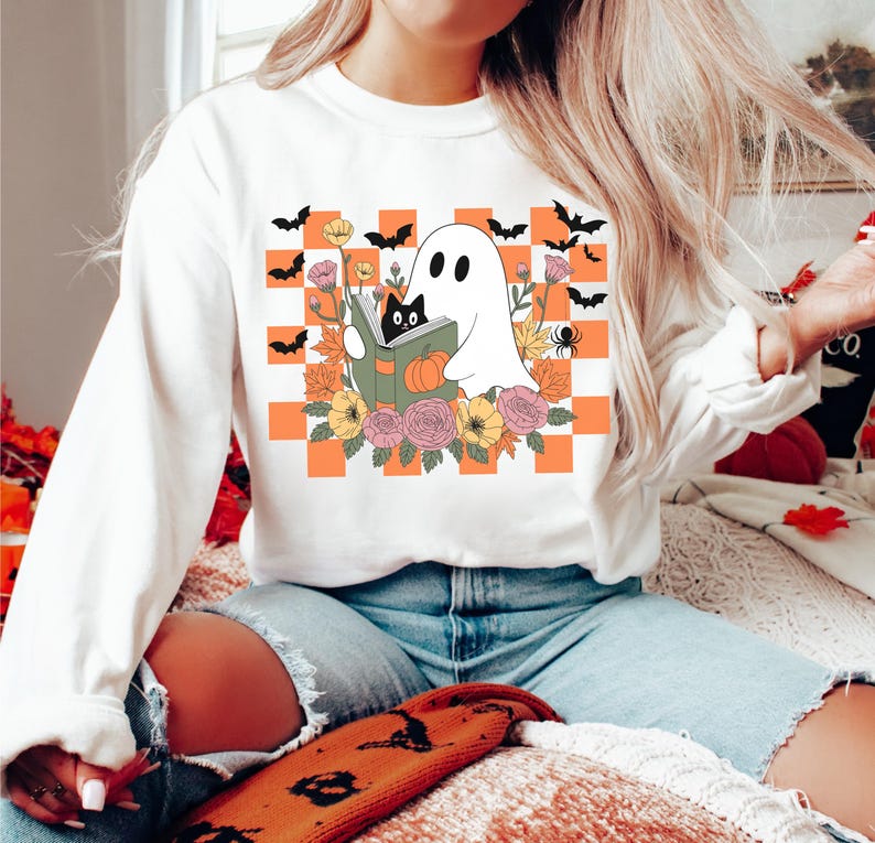 May include: White sweatshirt featuring a Halloween-themed graphic. The design includes a ghost reading a book with a black cat, surrounded by flowers, pumpkins, and bats, set against an orange and white checkered background.