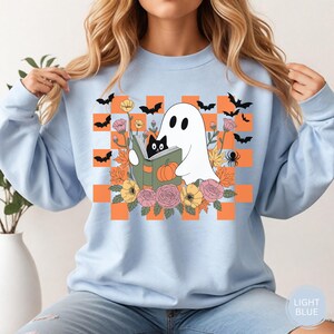 May include: Light blue sweatshirt with a Halloween-themed graphic. The design features a white ghost reading a book with a black cat, surrounded by flowers, bats, and a spider against an orange and white checkered background.