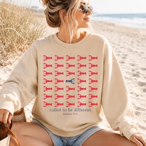 May include: A sand-colored crewneck sweatshirt featuring a grid of red lobster icons with one blue lobster in the center. Below the grid, the text reads "called to be different" in blue, followed by the smaller blue text "Romans 12:2".