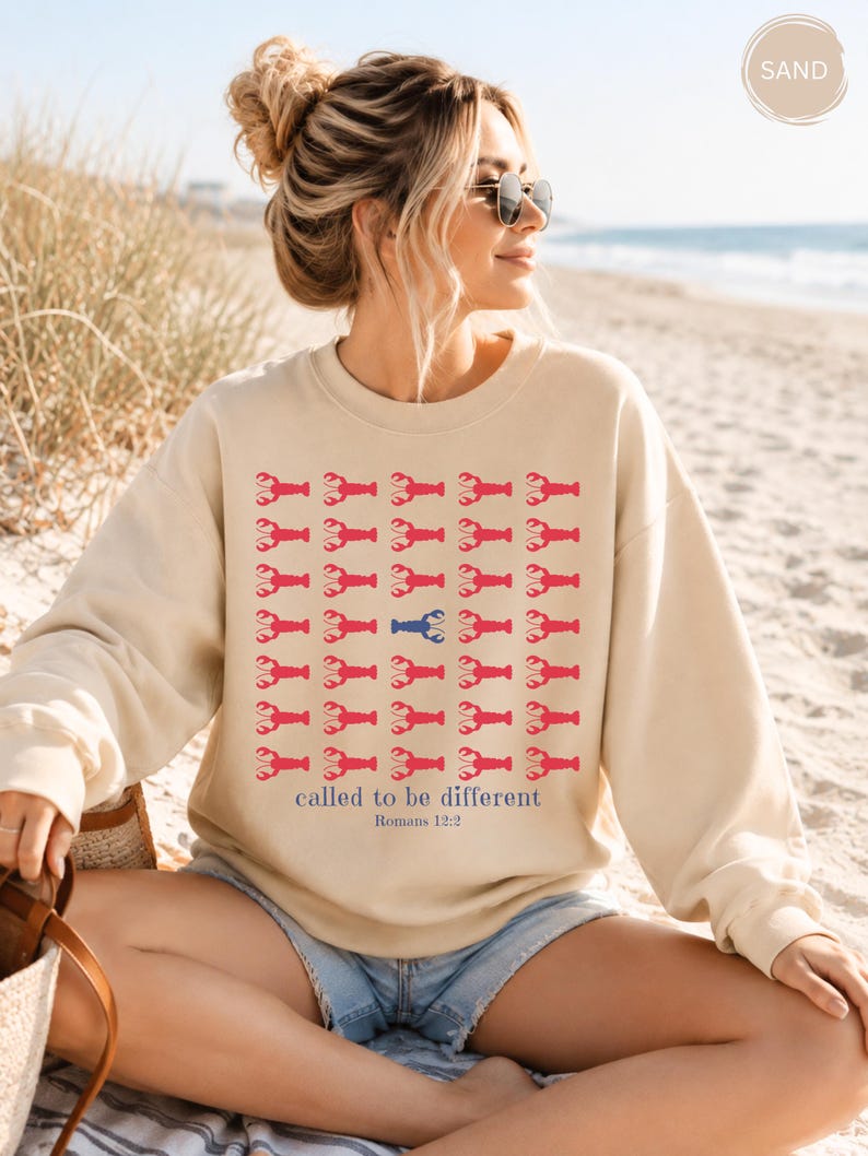 May include: A sand-colored crewneck sweatshirt featuring a grid of red lobster icons with one blue lobster in the center. Below the grid, the text reads "called to be different" in blue, followed by the smaller blue text "Romans 12:2".