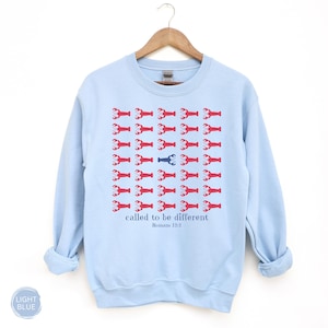 May include: A light blue crewneck sweatshirt featuring a grid of red lobster graphics with one central blue lobster. Below the grid, the text reads "called to be different" in blue, followed by the smaller blue text "Romans 12:2" centered below.