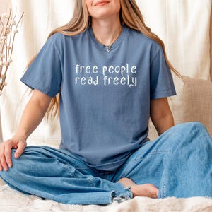 May include: A blue t-shirt with the text "free people read freely" in white script. The person is wearing blue jeans. A white vase with dried flowers is on the left. A beige pillow with buttons is on the right.