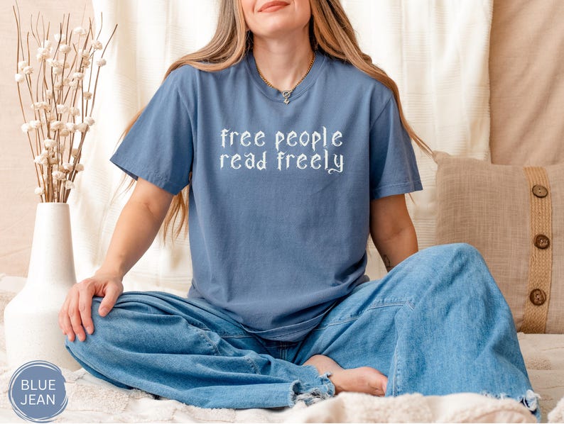 May include: A blue t-shirt with the text "free people read freely" in white script. The person is wearing blue jeans. A white vase with dried flowers is on the left. A beige pillow with buttons is on the right.