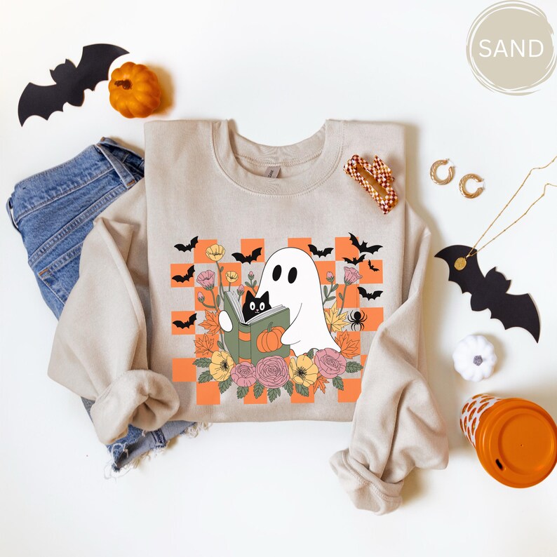 May include: Sand-colored sweatshirt featuring a Halloween-themed graphic. The design includes a white ghost reading a book with a black cat, surrounded by flowers, bats, and a spider. The background has an orange and white checkered pattern.