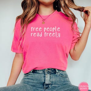 May include: A bright pink t-shirt with the text "free people read freely" in white script. The shirt is a short-sleeved crew neck style. The model is wearing blue jeans.