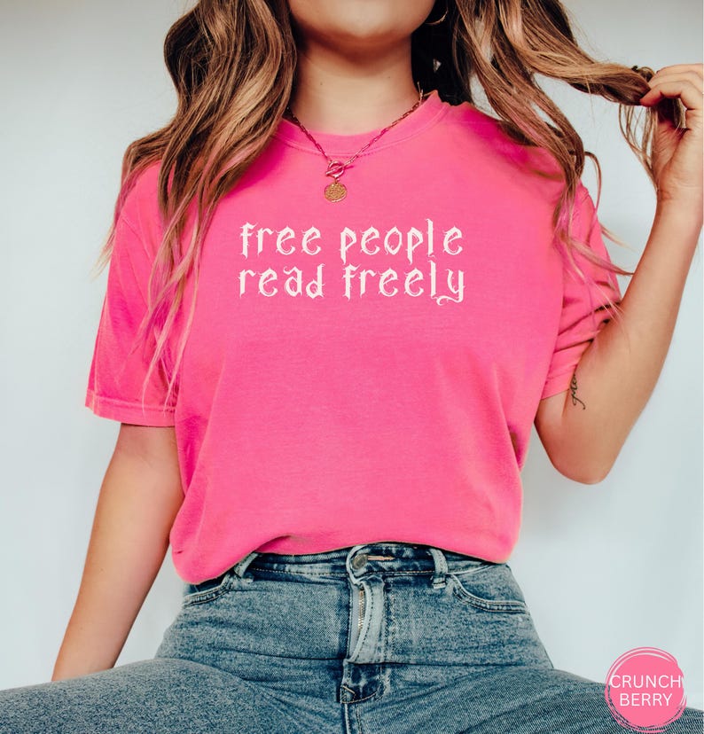 May include: A bright pink t-shirt with the text "free people read freely" in white script. The shirt is a short-sleeved crew neck style. The model is wearing blue jeans.
