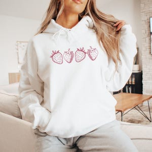 May include: White hoodie with a pink strawberry graphic design. The strawberries are arranged in a row across the front of the hoodie.