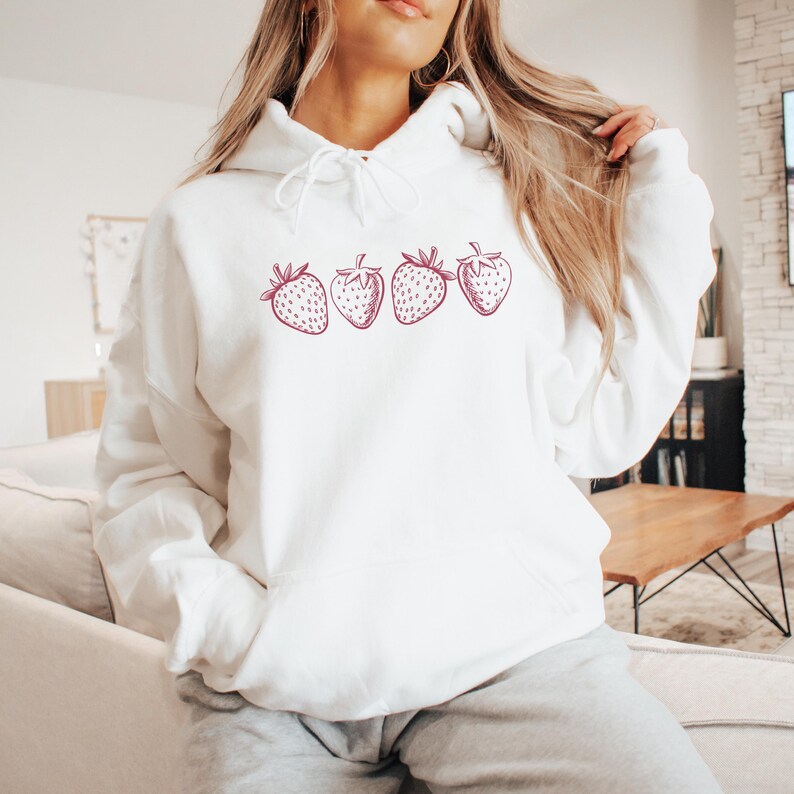 May include: White hoodie with a pink strawberry graphic design. The strawberries are arranged in a row across the front of the hoodie.