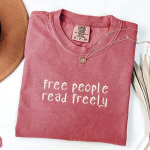 May include: A crimson-colored t-shirt with the text "free people read freely" in a decorative font. A silver necklace with a pendant rests on the shirt. The shirt is folded and placed on a white surface, with a brown hat and dried plant in the background.
