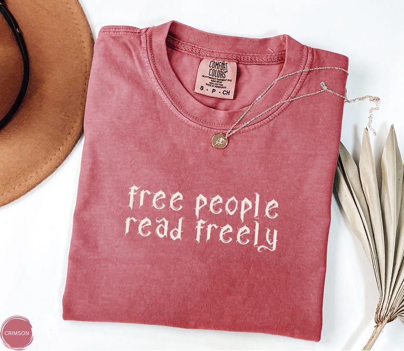 May include: A crimson-colored t-shirt with the text "free people read freely" in a decorative font. A silver necklace with a pendant rests on the shirt. The shirt is folded and placed on a white surface, with a brown hat and dried plant in the background.