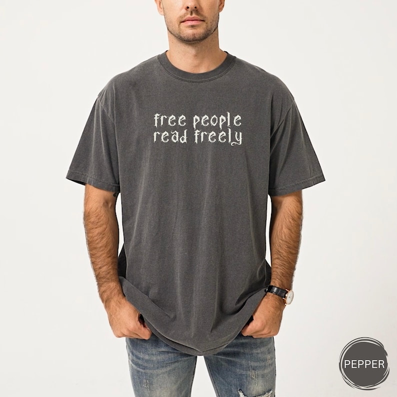 May include: Dark gray t-shirt with the text "free people read freely" in white script. The shirt is a short-sleeved, crew neck style. The model is wearing blue jeans and a watch.