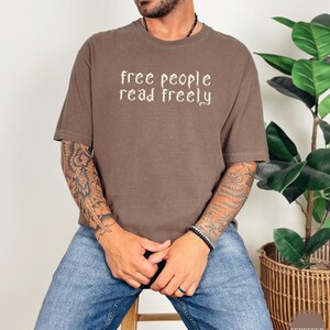 May include: Espresso-colored t-shirt with the text "free people read freely" in a decorative font. The shirt is a relaxed fit and is paired with blue jeans. The image is taken indoors.