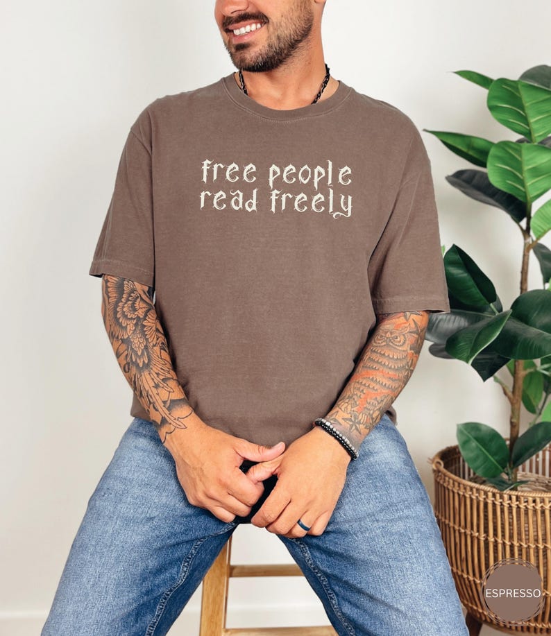 May include: Espresso-colored t-shirt with the text "free people read freely" in a decorative font. The shirt is a relaxed fit and is paired with blue jeans. The image is taken indoors.