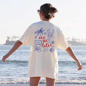 Puede incluir: Una camiseta blanca con un estampado gr&aacute;fico azul y rojo que dice "sea ya later" con una langosta, palmeras y otras im&aacute;genes de tem&aacute;tica playera.