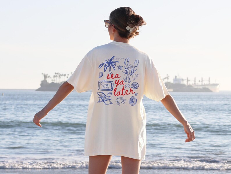 Puede incluir: Una camiseta blanca con un estampado gr&aacute;fico azul y rojo que dice "sea ya later" con una langosta, palmeras y otras im&aacute;genes de tem&aacute;tica playera.