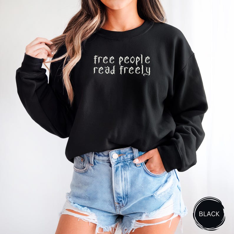 Może przedstawiać: Czarna bluza z białym napisem "free people read freely" w gotyckiej czcionce. Top z długim rękawem jest połączony z dżinsowymi szortami. Czarne koło ze słowem "BLACK" znajduje się w prawym dolnym rogu.