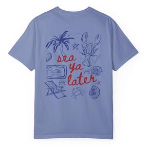 Puede incluir: Una camiseta azul claro con un estampado gr&aacute;fico azul y rojo de una langosta, una palmera, conchas marinas y una lata de sardinas. El texto de la camiseta dice "sea ya later".