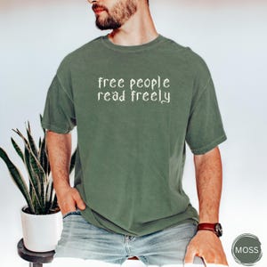 May include: Olive green t-shirt with the text "free people read freely" in white. The shirt has short sleeves and a crew neck. The shirt is made of a soft, comfortable material.