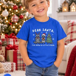 Puede incluir: Una camiseta de manga corta color azul real con un gr&aacute;fico de tres mapaches y el texto "DEAR SANTA, I'VE BEEN A LITTLE FERAL". Los mapaches est&aacute;n decorados con astas de reno, un gorro de Pap&aacute; Noel, una pajarita roja y luces de Navidad.