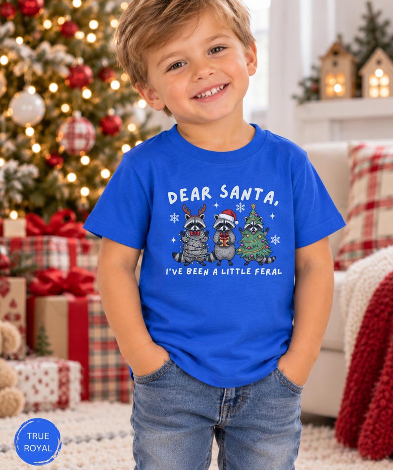 Puede incluir: Una camiseta de manga corta color azul real con un gr&aacute;fico de tres mapaches y el texto "DEAR SANTA, I'VE BEEN A LITTLE FERAL". Los mapaches est&aacute;n decorados con astas de reno, un gorro de Pap&aacute; Noel, una pajarita roja y luces de Navidad.