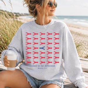 May include: A light gray crewneck sweatshirt featuring a repeating grid pattern of red lobster icons, with one blue lobster icon in the center. Below the grid, the text reads "called to be different" in blue, followed by "Romans 12:2" in smaller blue text.