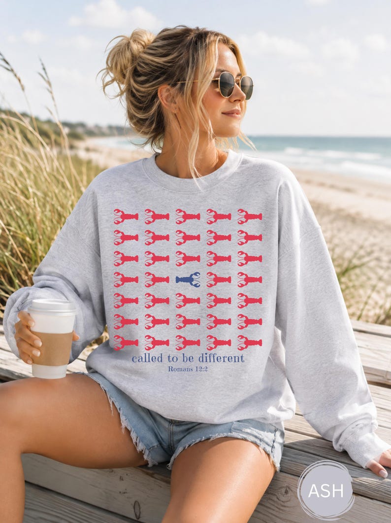 May include: A light gray crewneck sweatshirt featuring a repeating grid pattern of red lobster icons, with one blue lobster icon in the center. Below the grid, the text reads "called to be different" in blue, followed by "Romans 12:2" in smaller blue text.
