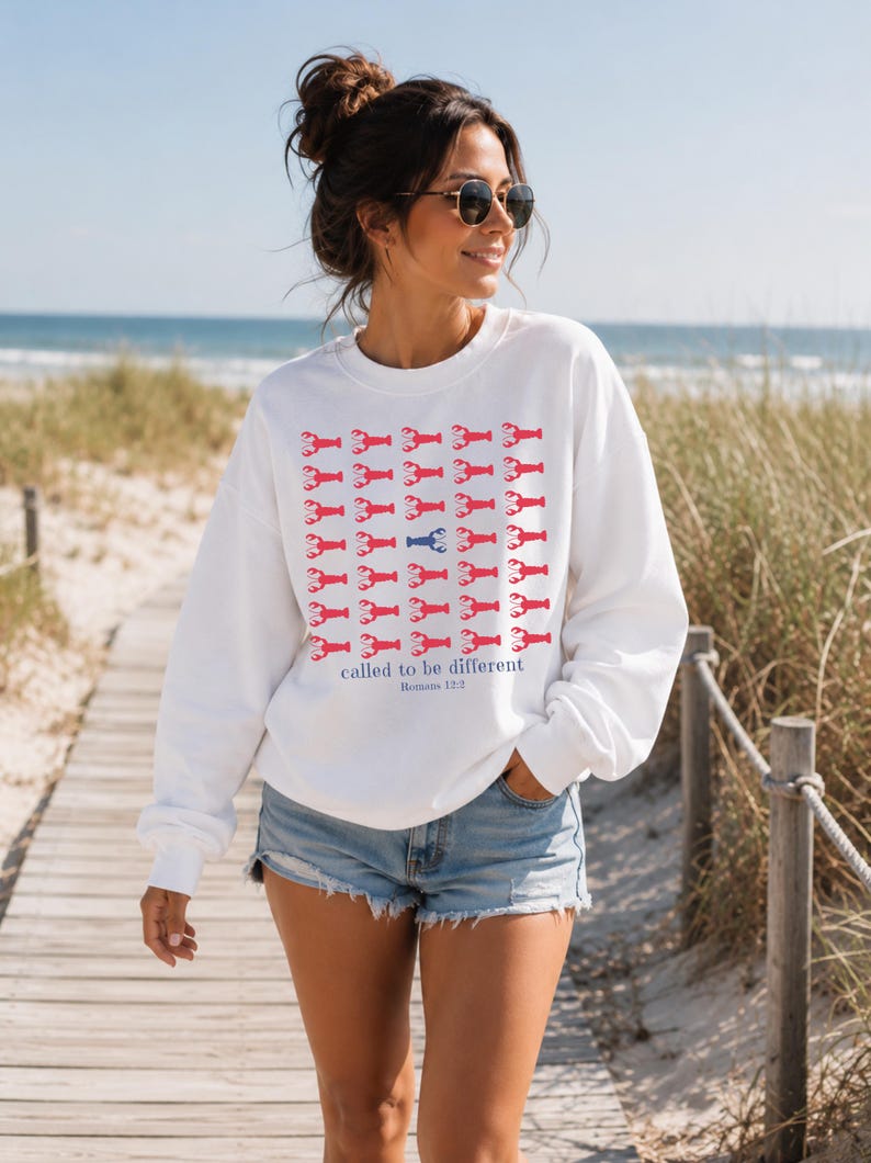 May include: A white long-sleeve crewneck sweatshirt featuring a grid pattern of small red lobster illustrations with one blue lobster in the center. Below the grid, the text reads "called to be different" followed by "Romans 12:2" in blue lettering.