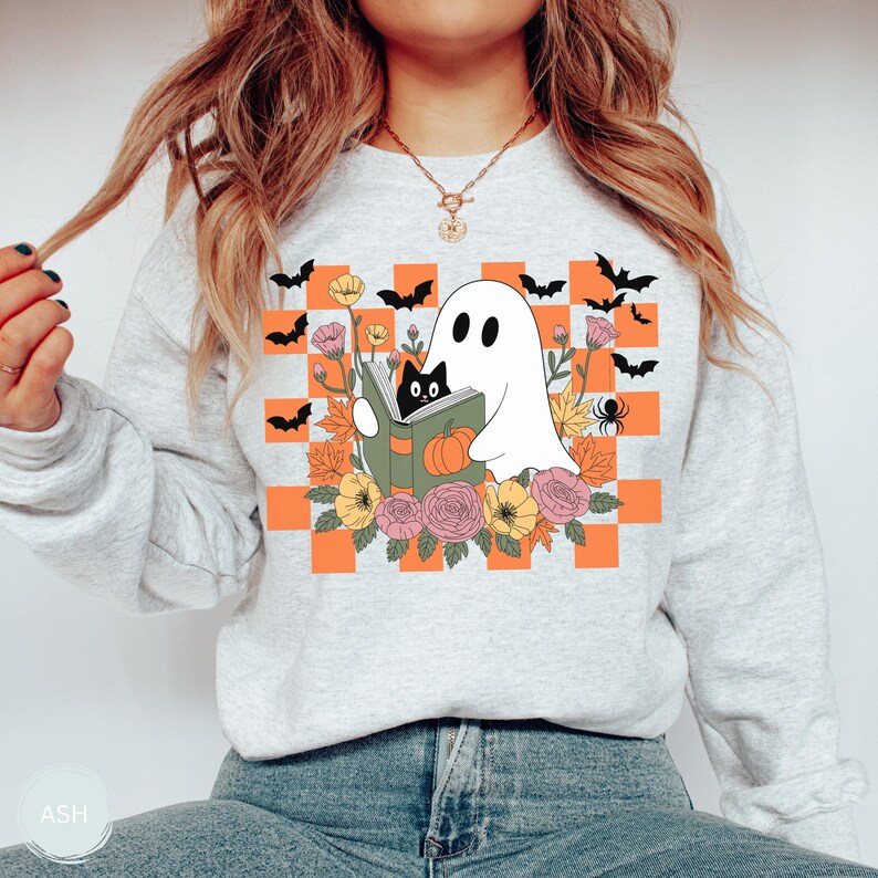 May include: A light gray sweatshirt featuring a Halloween-themed graphic. The design includes a white ghost reading a book with a black cat, surrounded by flowers, bats, and a spider, set against an orange and white checkered background.