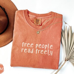 May include: A coral-colored t-shirt with the phrase "free people read freely" in white script. A gold necklace with a pendant rests on the shirt. A brown hat and dried palm leaves are also in the image.